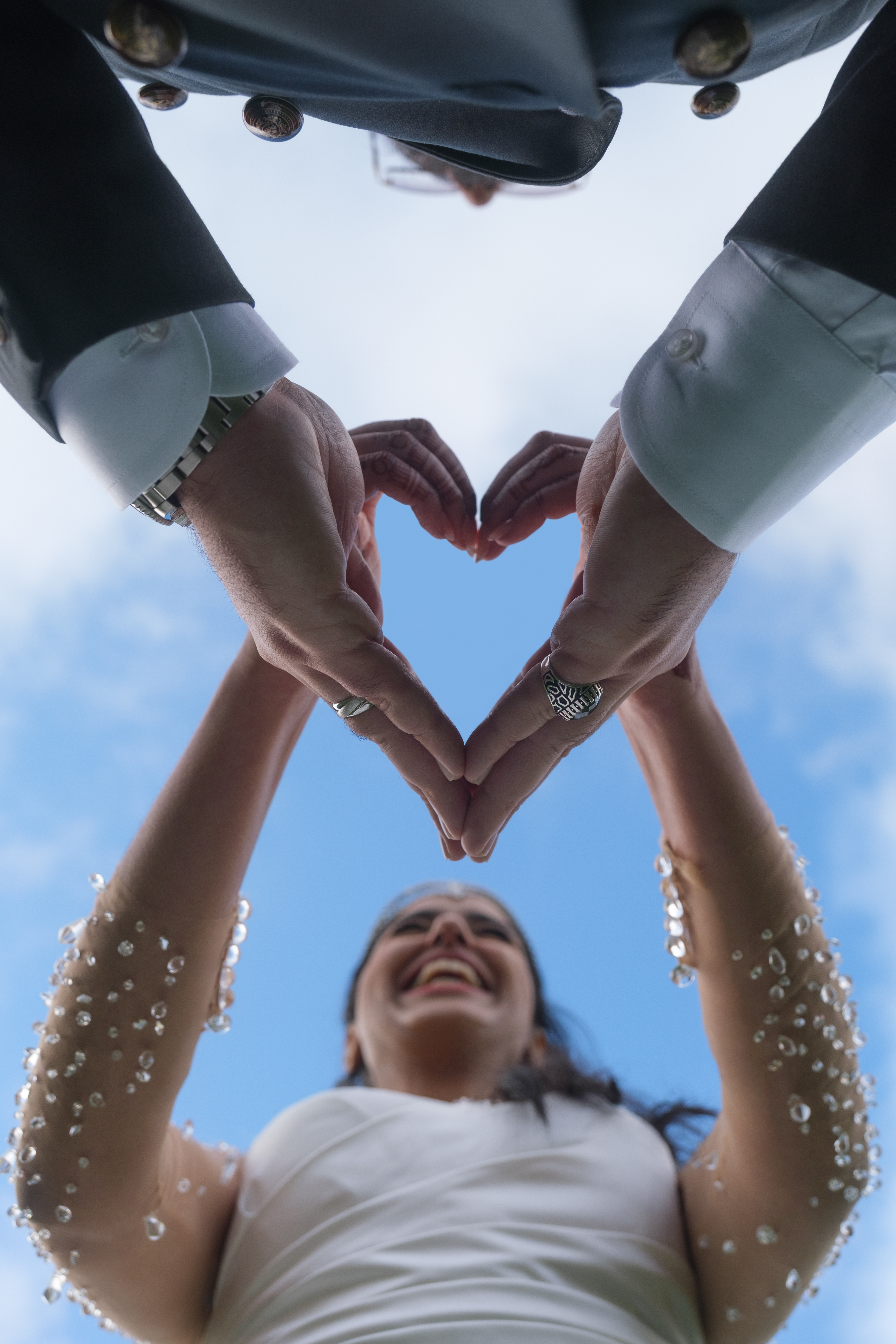 Photo de mariage vue en contre-plongée, mains formant un cœur bleu sous un ciel clair, mariée souriante.