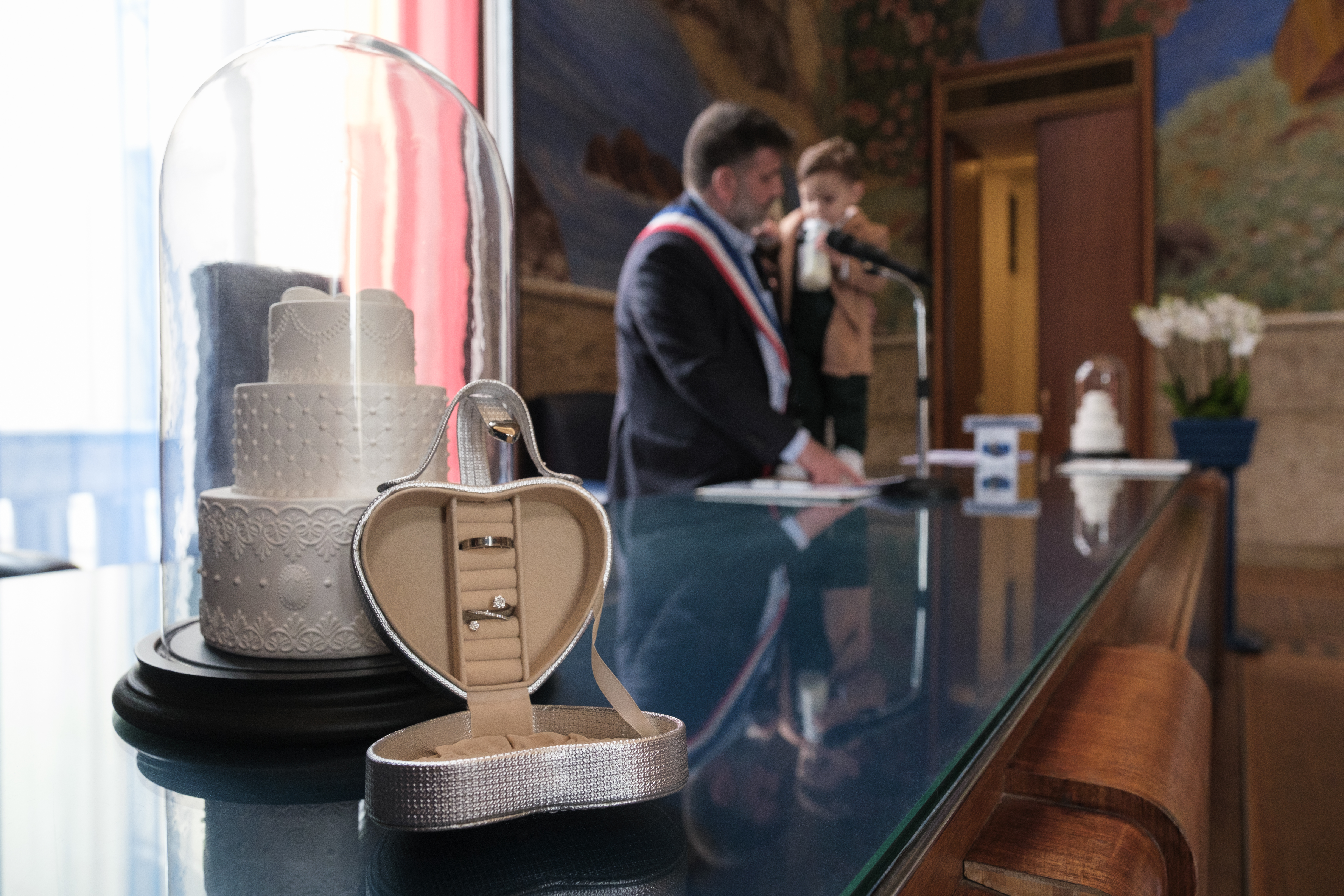 Écrin à bijoux en forme de cœur contenant des alliances de mariage, posé sur une table en verre, avec en arrière-plan un homme tenant un enfant lors d’une cérémonie.