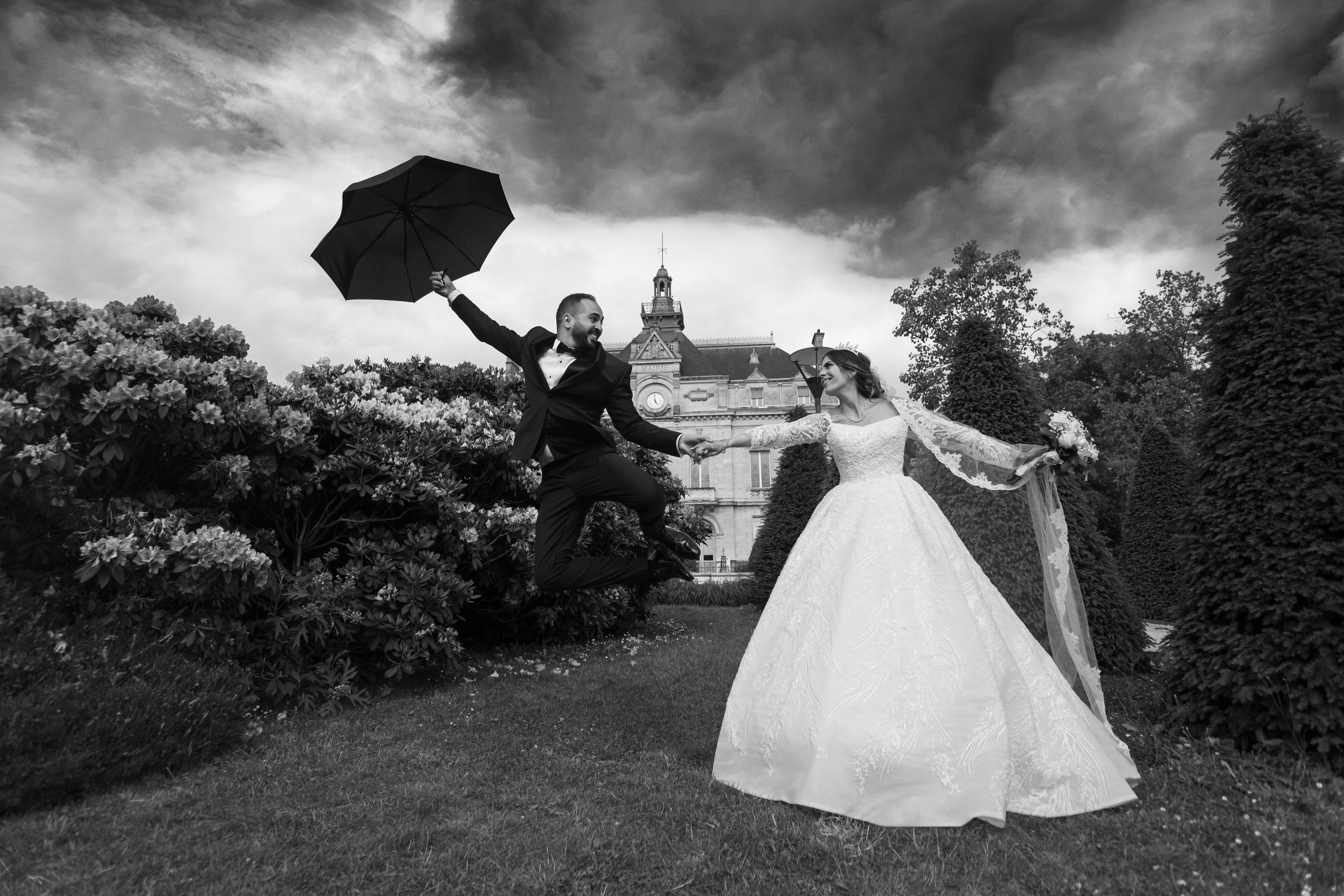 Mariage en noir et blanc, marié sautant avec un parapluie, mariée en robe blanche dans un jardin sous un ciel orageux, photo pleine d’émotion et de joie.